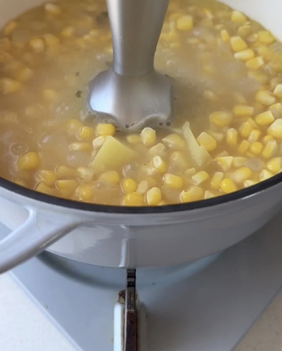 Ein Stabmixer püriert die köchelnden Maiskörner und Süßkartoffelstücke im Topf zu einer glatten Flüssigkeit.