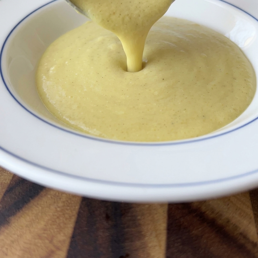 Dickflüssige, cremige gelbe Maissuppe wird aus einer Schöpfkelle in eine weiße Schüssel mit blauem Rand gefüllt.