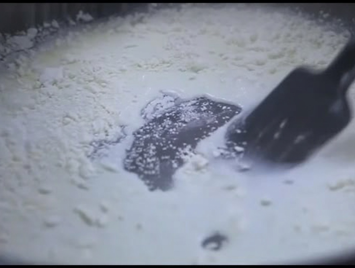 Ein schwarzer Silikonspatel rührt eine dicke, weiße Mehl-Milch-Mischung in einer Pfanne um.