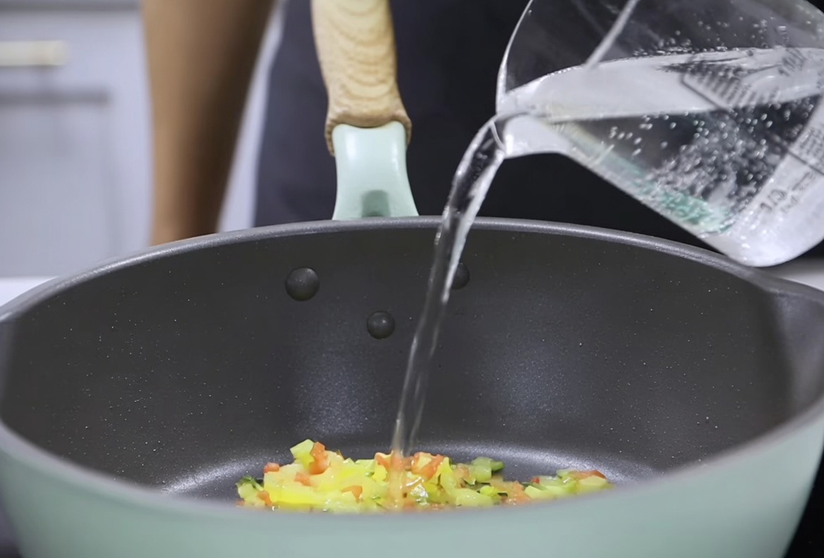 Wasser wird aus einem Messbecher in eine Pfanne zum Gemüse gegossen, um die cremige Mischung zu vervollständigen.