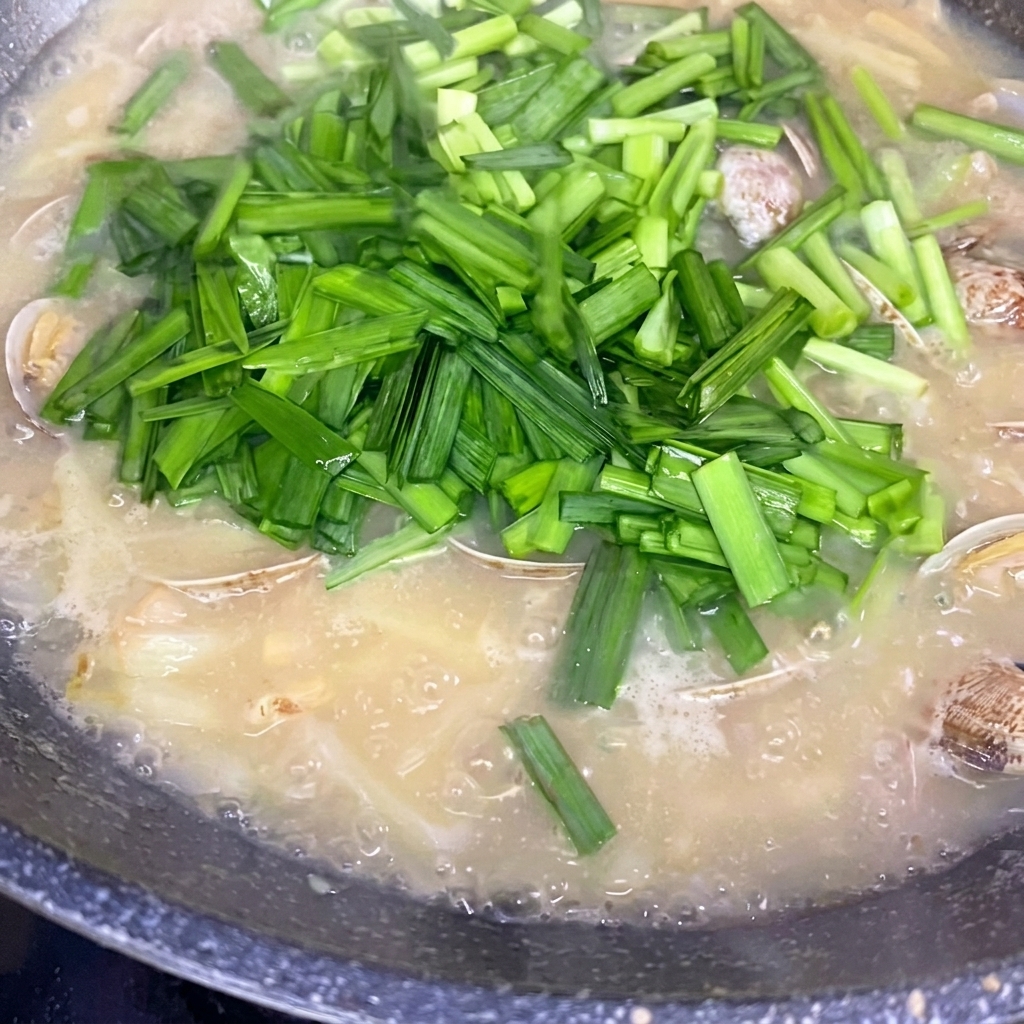 Frisch gehackter chinesischer Schnittlauch wird über die köchelnde Kartoffel-Muschel-Suppe gestreut.