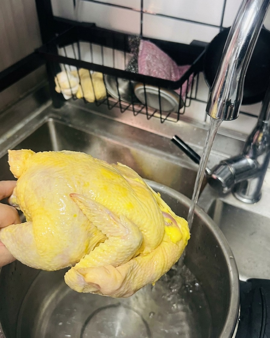 Ein ganzes, rohes gelbes Huhn wird unter fließendem Wasser gewaschen, bevor es in einen Topf mit heißem Wasser kommt.