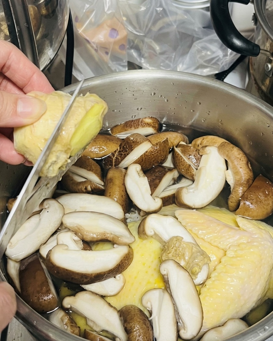 Jemand schneidet ein Stück frischen Ingwer direkt über einem Topf mit Huhn und Pilzen.