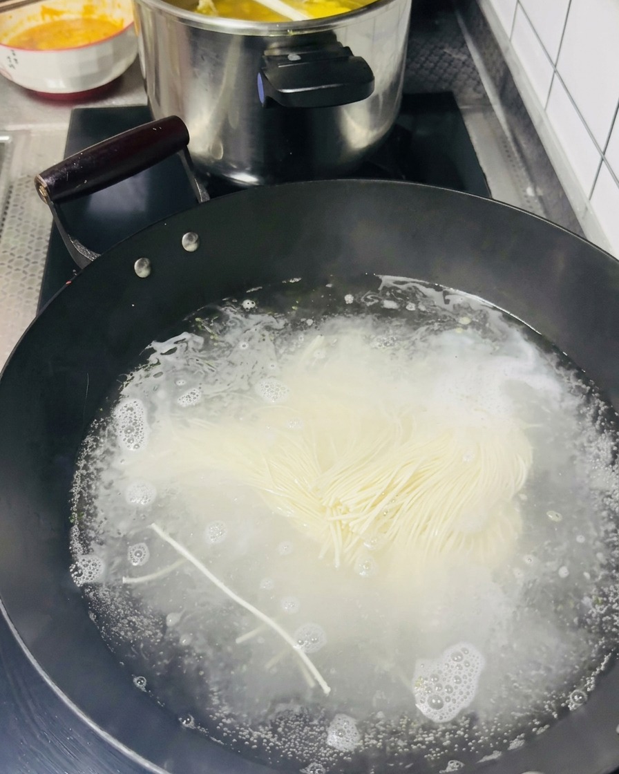Ein Bündel dünner, roher heller Nudeln wird in einen schwarzen Wok mit sprudelnd kochendem Wasser gegeben.