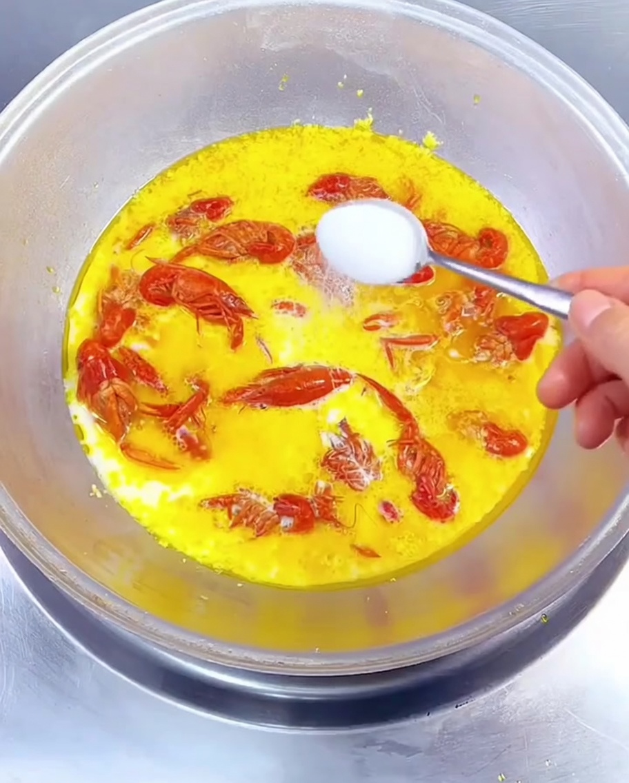 Nahaufnahme eines Löffels, der weiße Gewürze in einen köchelnden Wok mit goldener Milch-Sauce und roten Flusskrebsen gibt.