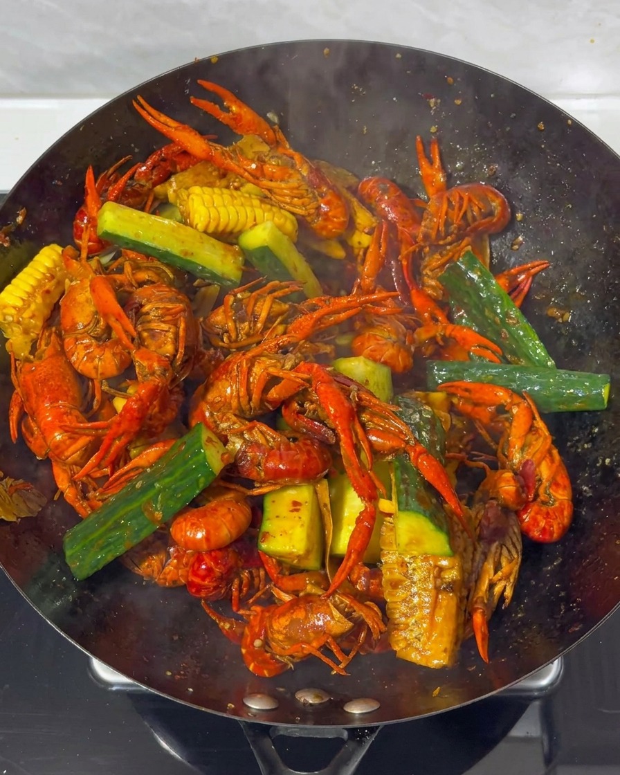 Nahaufnahme von Flusskrebsen, Mais und Gurkenstücken, die in einem großen Wok mit einer dicken, dunkelroten Sauce vermengt werden.
