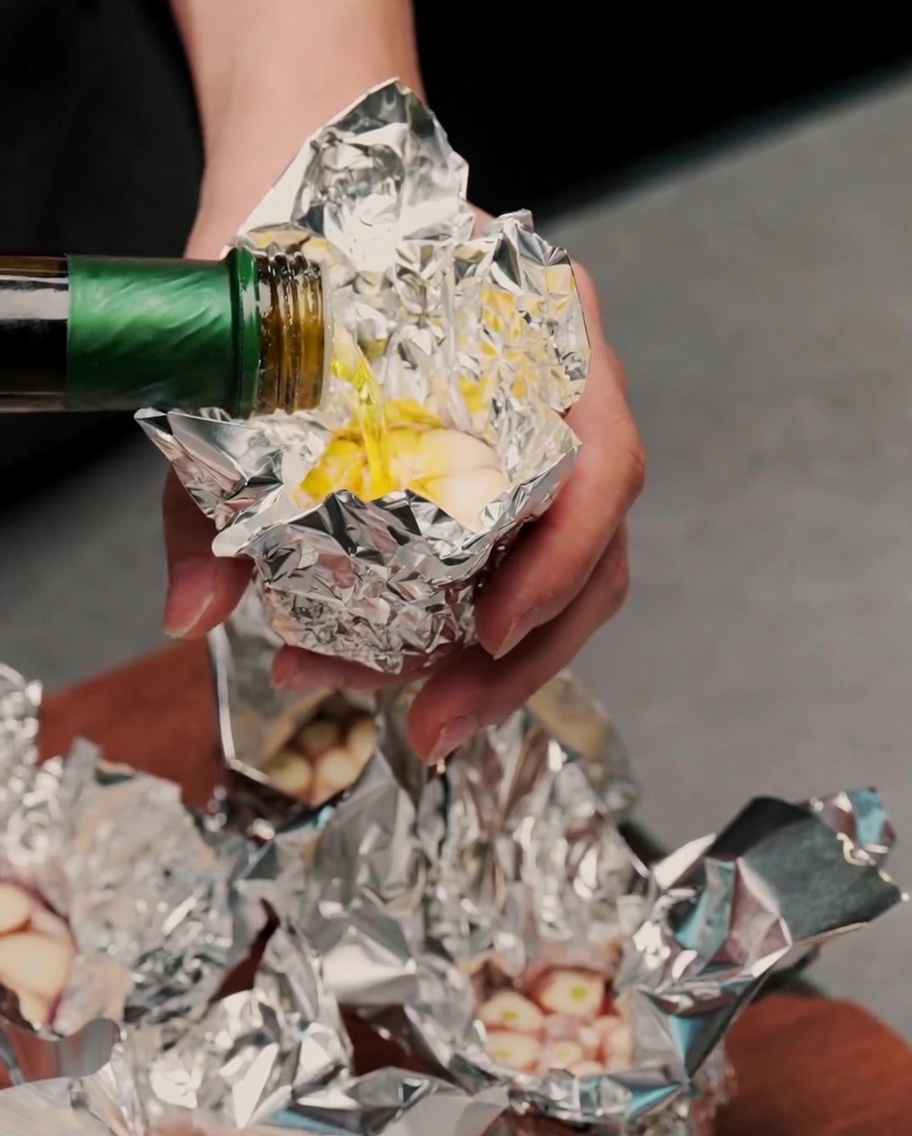 Olivenöl wird aus einer grünen Flasche auf die freiliegenden Knoblauchzehen in Alufolie gegossen.