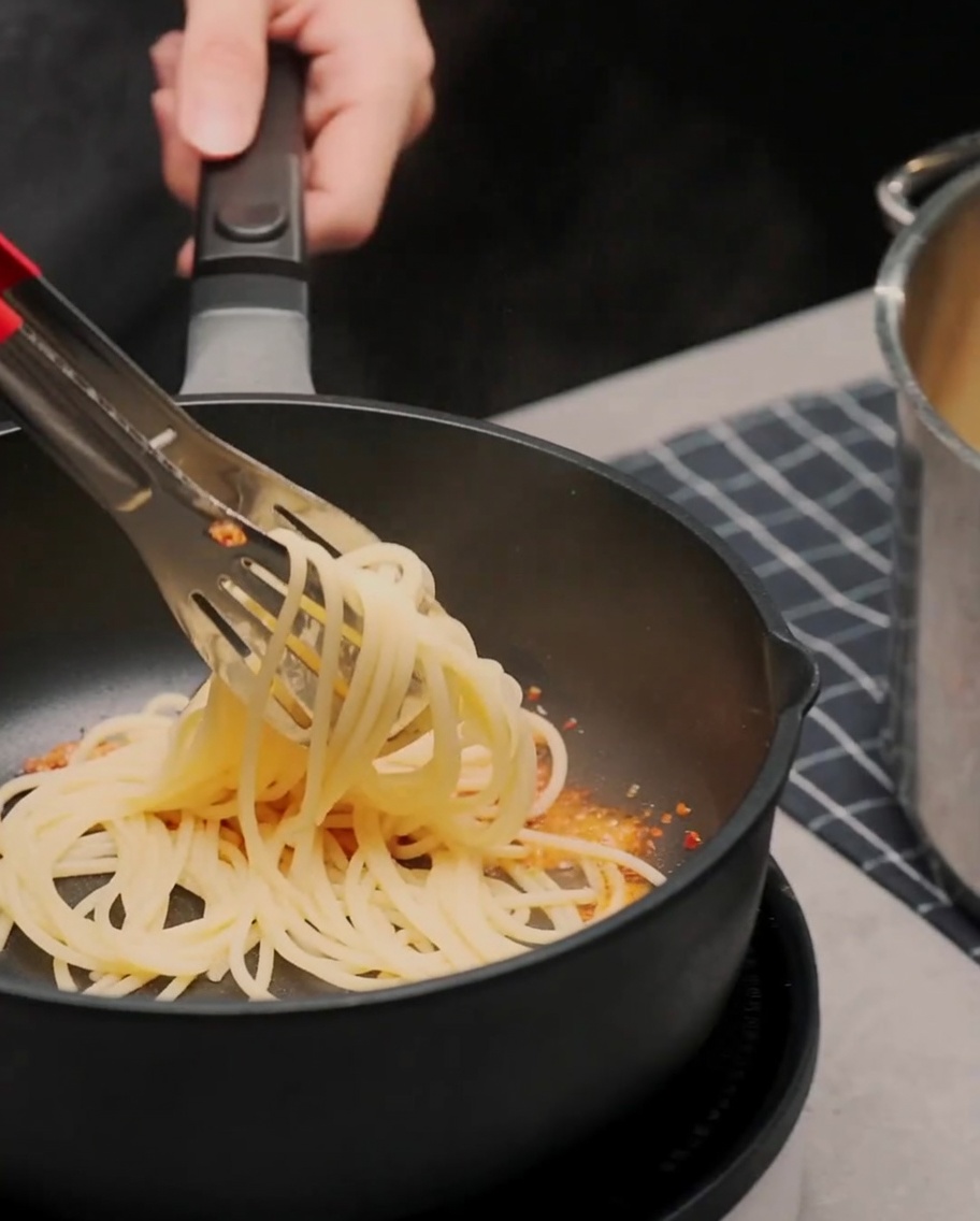 Spaghetti werden mit einer Zange in der Pfanne mit der Sauce vermengt.