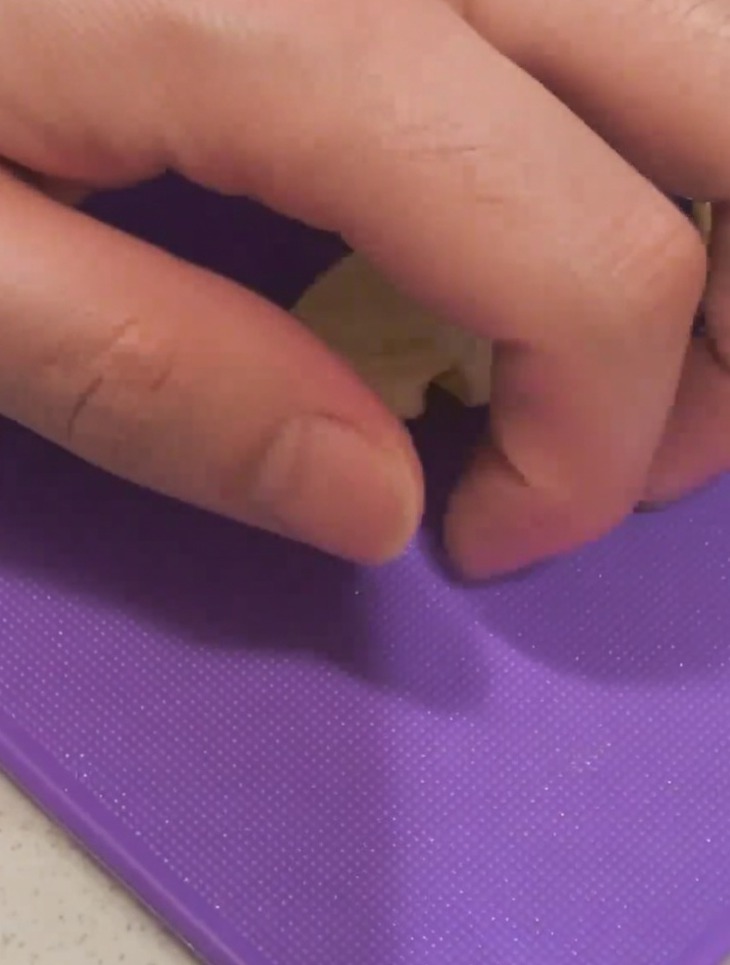 Nahaufnahme einer Hand, die mit der flachen Seite eines Messers eine Knoblauchzehe auf einem lila Schneidebrett zerdrückt.