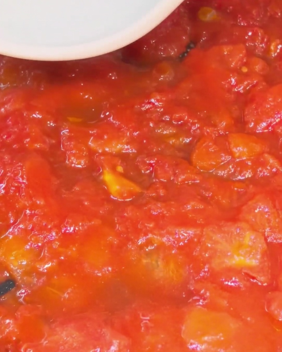 Nahaufnahme eines weißen Silikonspatels, der rote Tomatenstücke in einer Pfanne zu einer dicken, glatten Soße zerdrückt.