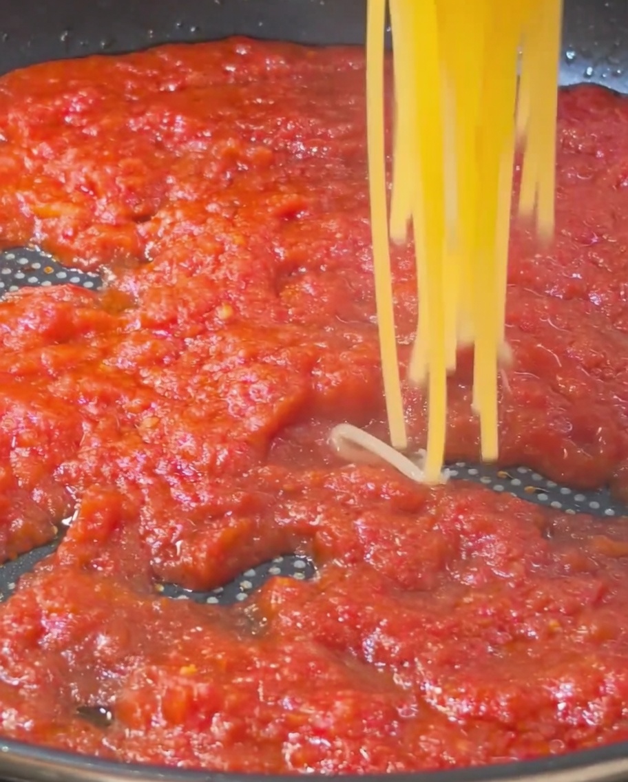 Eine Portion gekochter goldener Spaghetti wird angehoben und in eine Pfanne mit einer reichhaltigen, roten Tomatensoße gegeben.