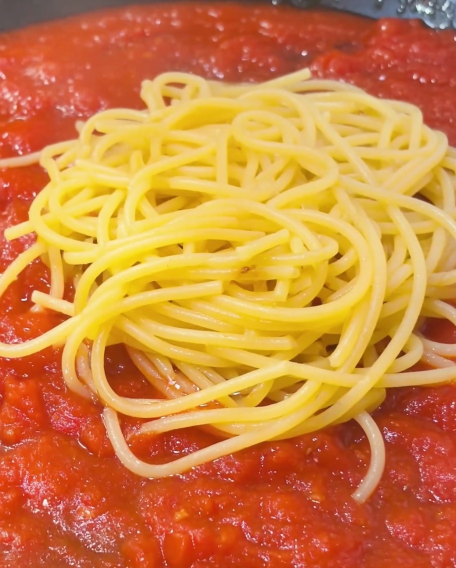Ein Löffel stärkehaltiges Nudelwasser wird über einen Berg Spaghetti und Tomatensoße in einer Pfanne geträufelt.