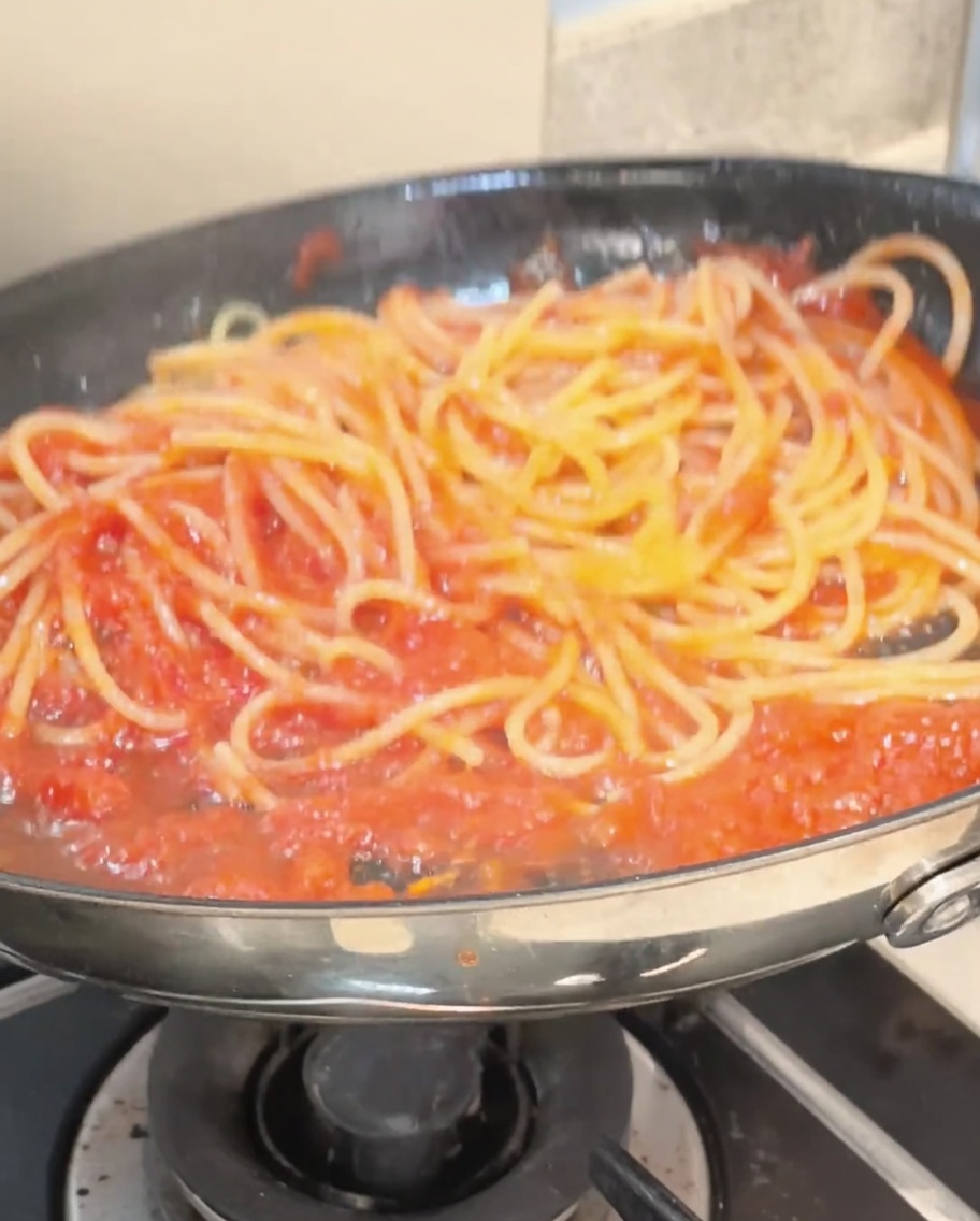 Spaghetti werden mit einer Zange in einer Pfanne geschwenkt und sind vollständig mit einer leuchtend roten Tomatensoße überzogen.