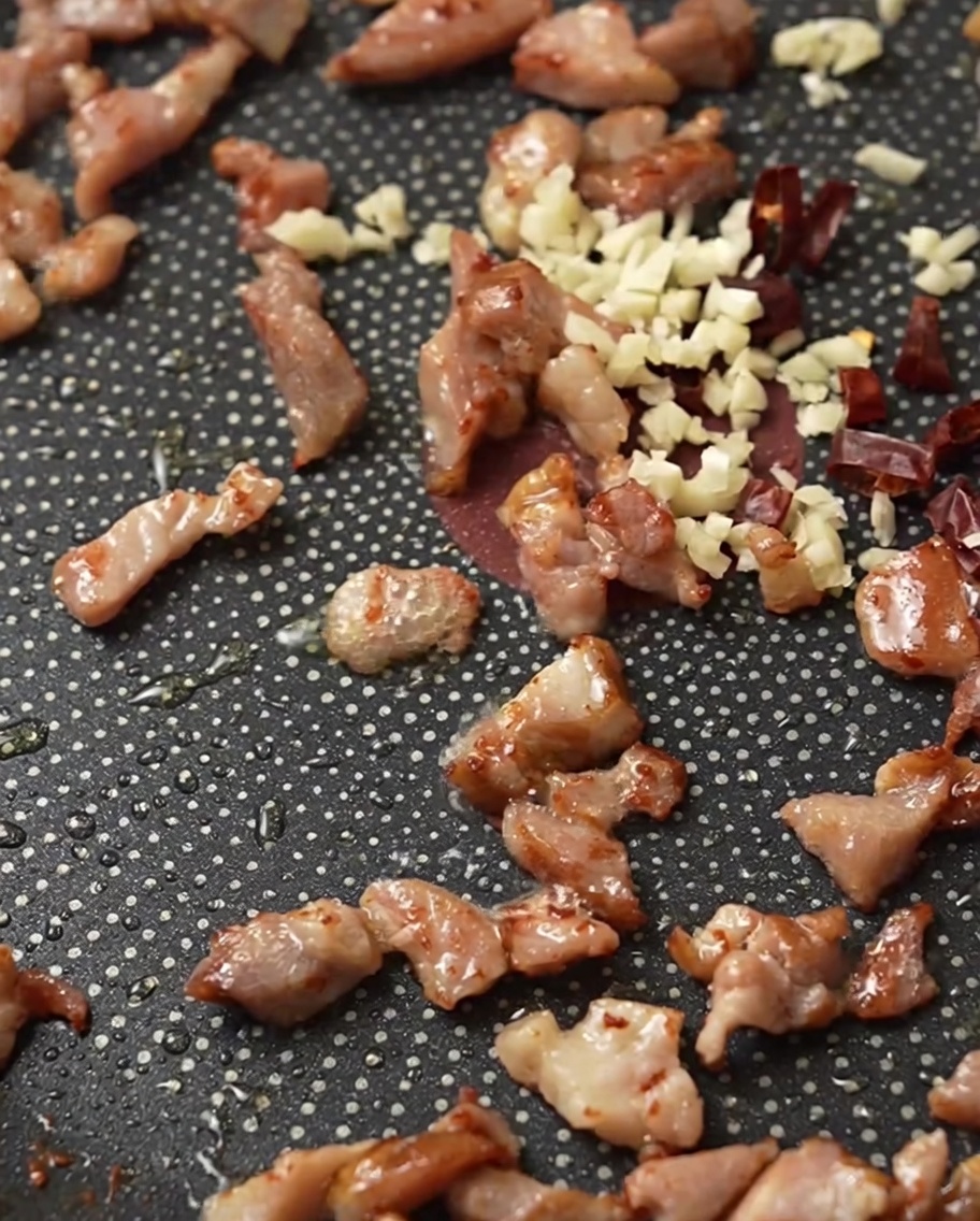 Knusprige Speckwürfel, gehackter Knoblauch und Chilis brutzeln zusammen in einer Bratpfanne.
