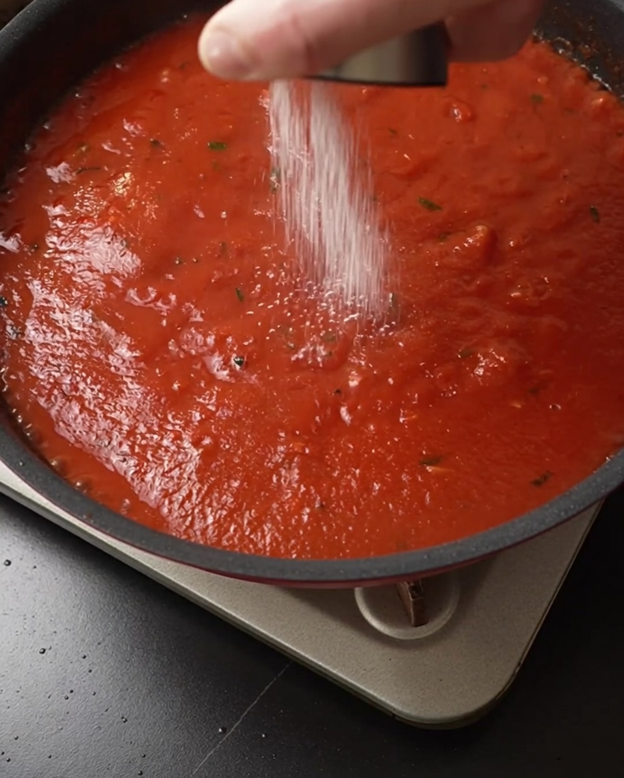Salz wird aus einem Streuer über eine köchelnde, tiefrote Tomatensoße gestreut.