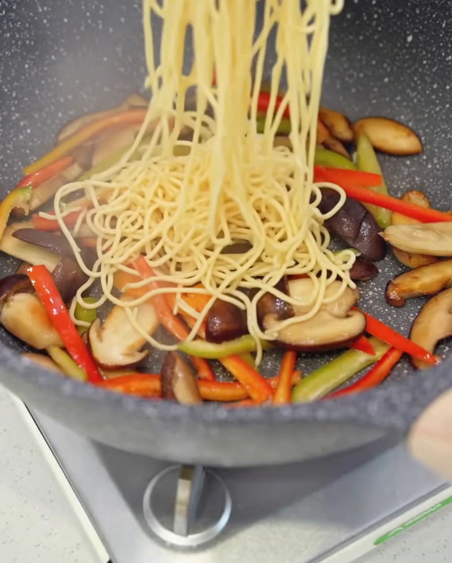 Gekochte Spaghetti werden in eine Pfanne mit gebratenen Pilzen und bunter Paprika gegeben.