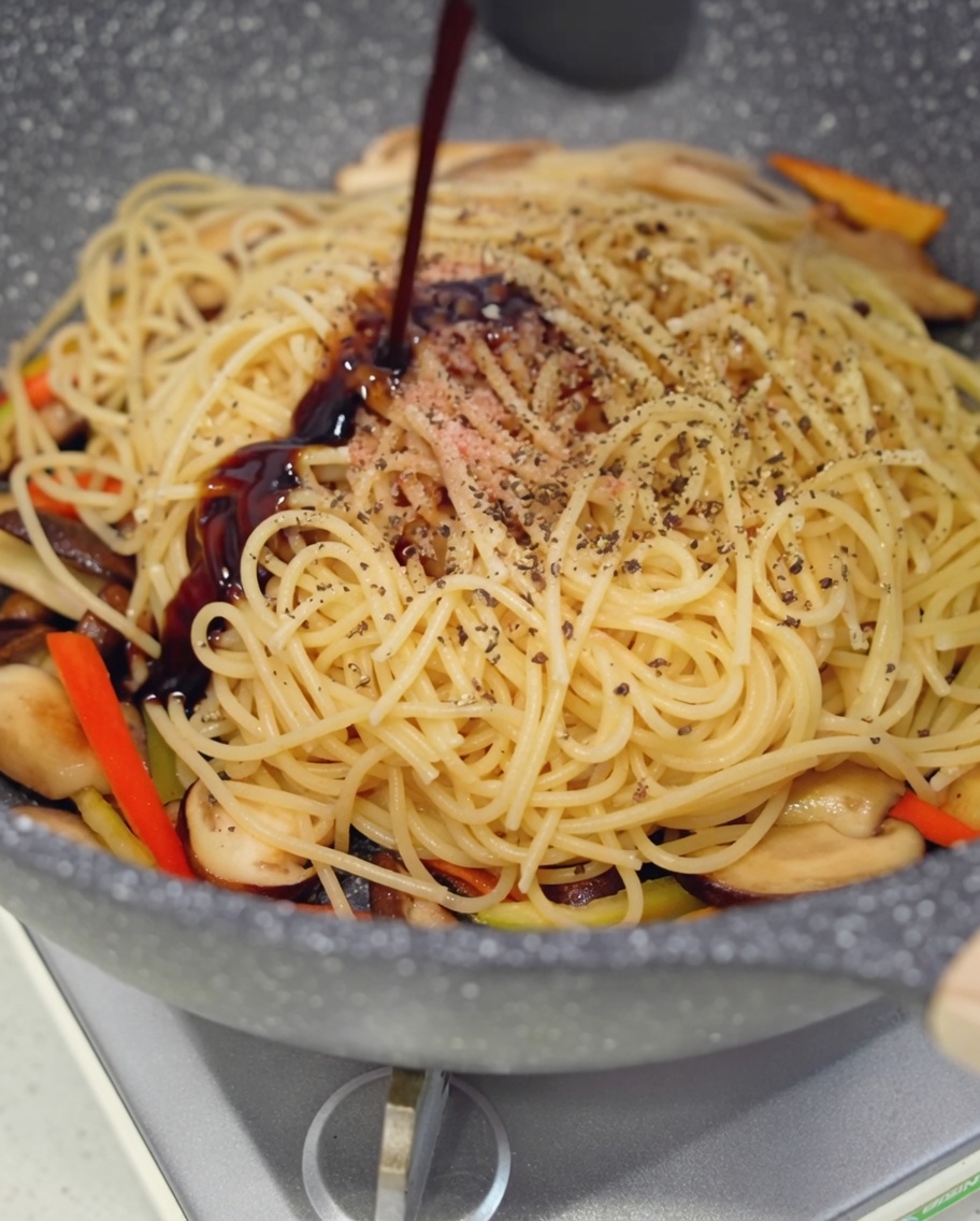 Dunkle vegane Austernsauce wird über die Nudel-Pilz-Mischung in der Pfanne geträufelt.