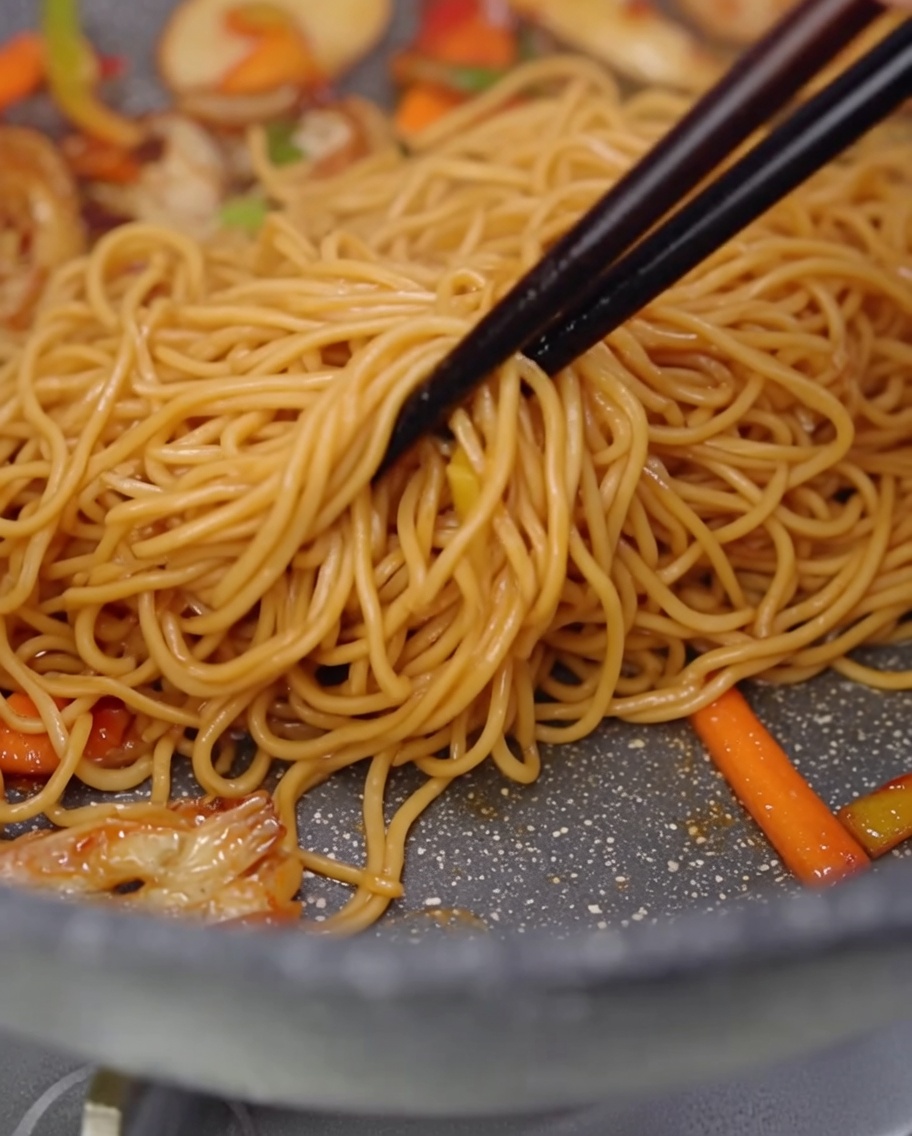 Eine Nahaufnahme von Spaghetti, geschnittenen Pilzen und bunten Paprikastreifen, die in einer Pfanne geschwenkt werden.