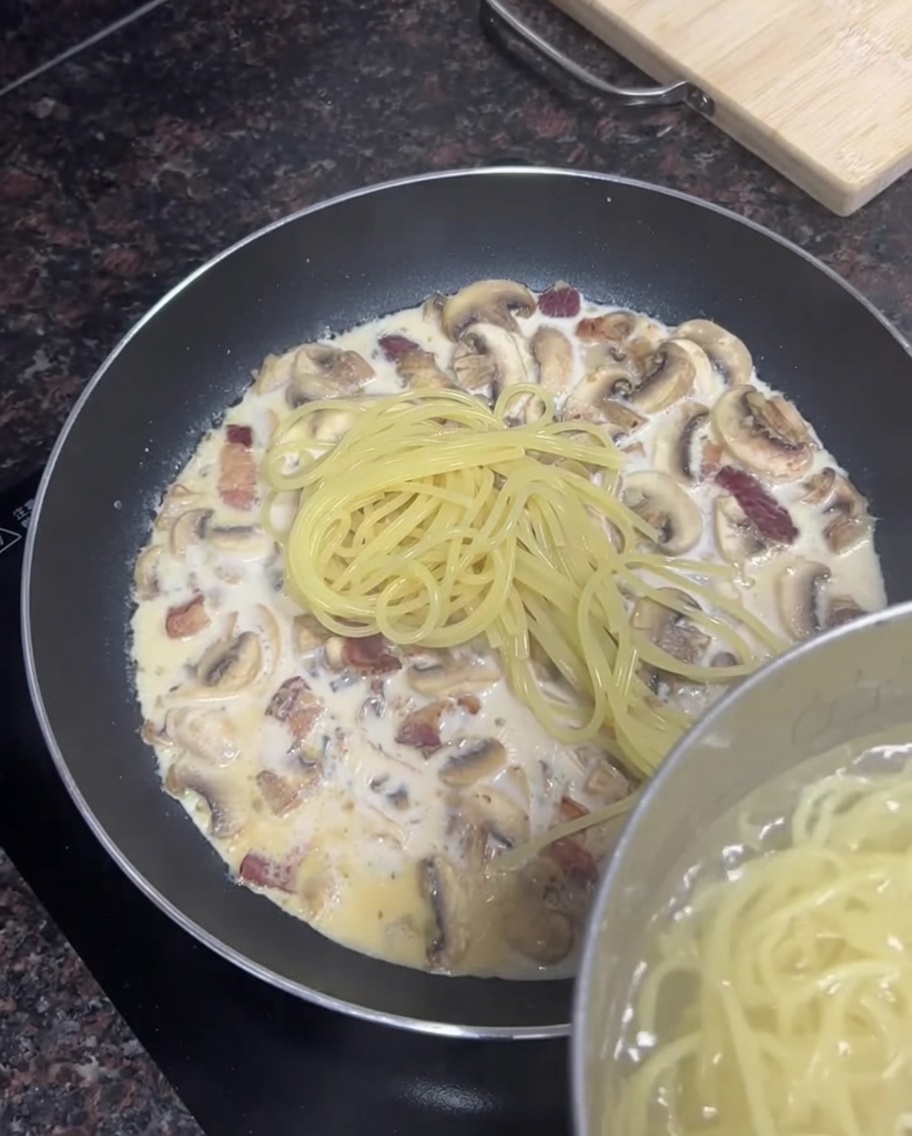 Gekochte Spaghetti werden aus dem Topf in die Pfanne mit cremiger Speck-Pilz-Sauce gegeben.