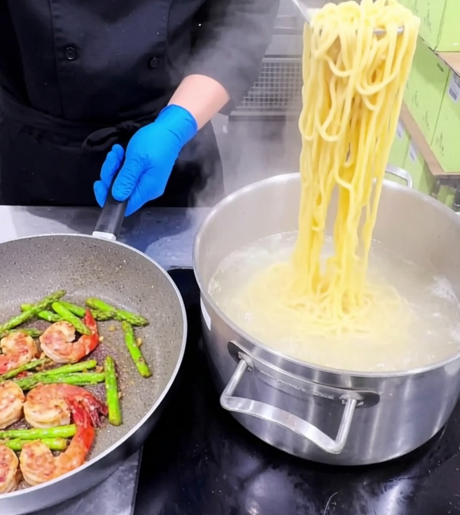 Pasta wird in die Pfanne mit Garnelen, Spargel und Knoblauch gegeben.