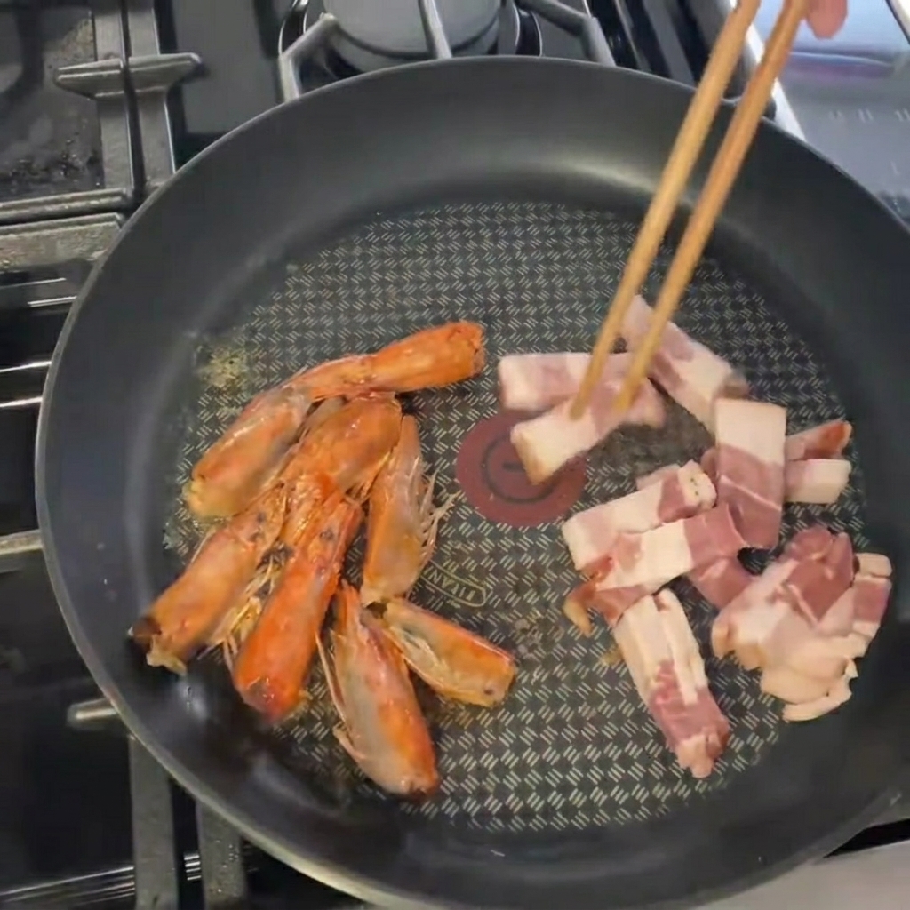 Speckstücke werden in die Pfanne zu den gebratenen Garnelenköpfen gegeben.