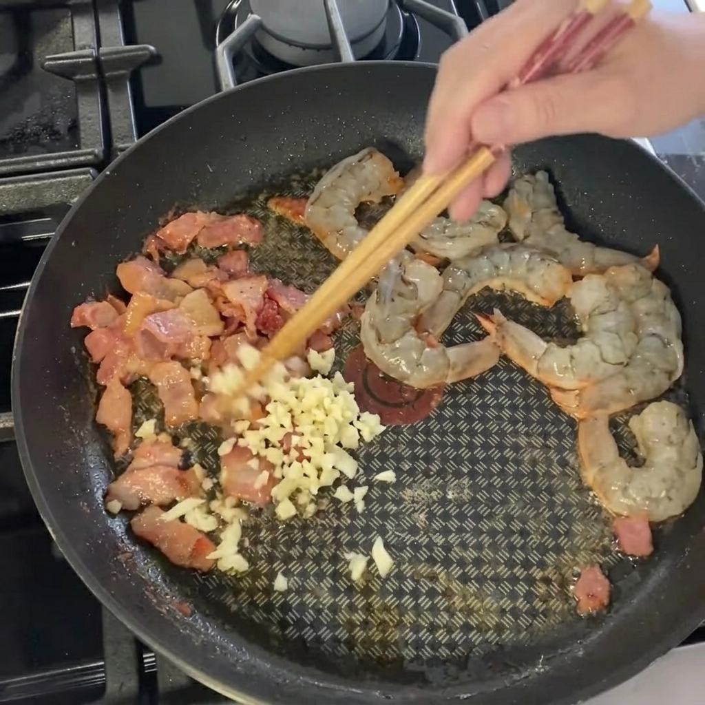 Fein gehackter Knoblauch wird über die Garnelen-Speck-Mischung gestreut.
