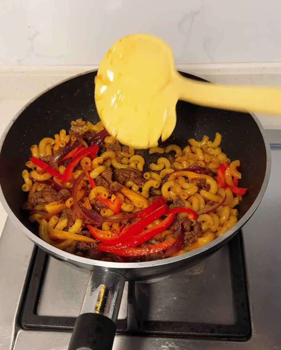 Eine Scheibe Käse liegt auf der Makkaroni-Mischung im Wok, während schwarzes Pfefferpulver darüber gestreut wird.