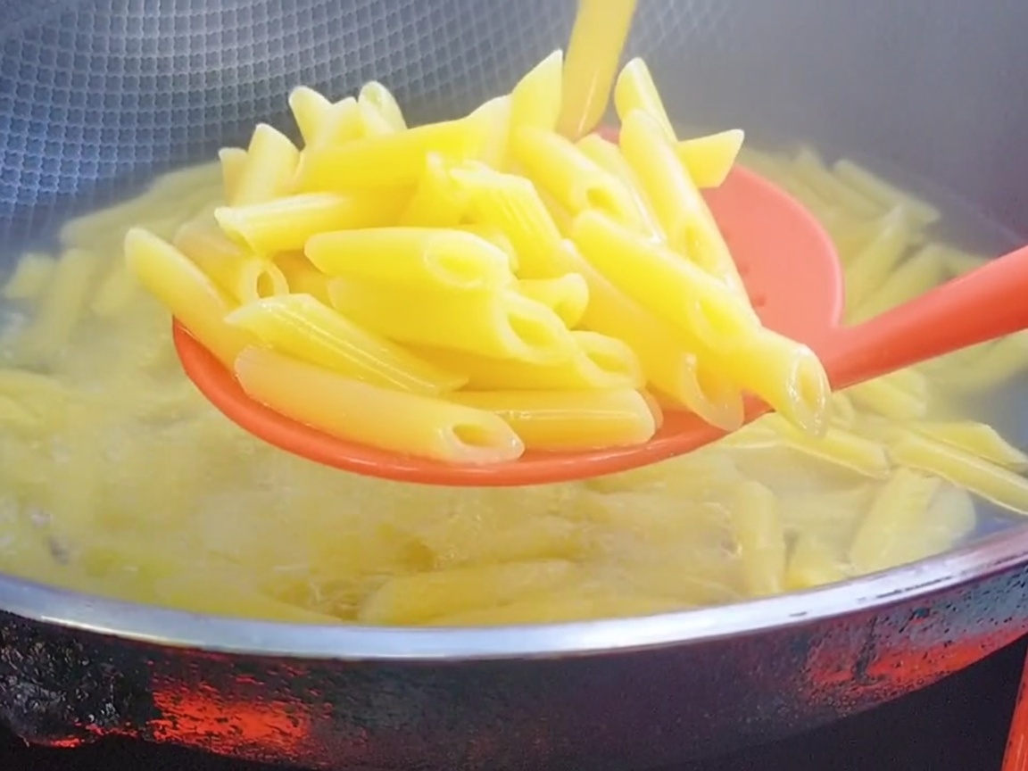 Ein roter Schaumlöffel aus Silikon hebt gekochte Penne aus dem kochenden Wasser.