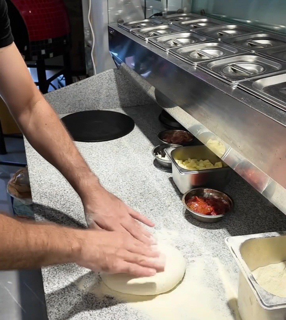 Hände, die eine Pizzateigkugel auf einer bemehlten Arbeitsfläche ausbreiten.