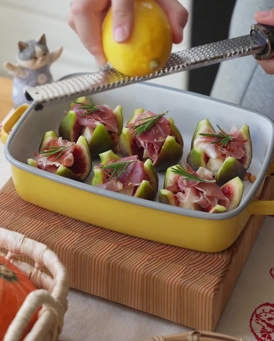 Frische Zitrone, deren Schale über ein Blech mit Schinken-gefüllten Feigen gerieben wird.