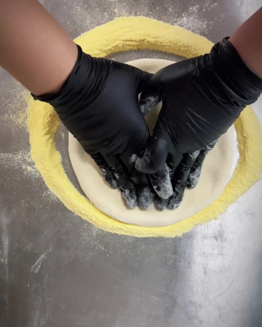 Hände in schwarzen Handschuhen drücken die Mitte des Pizzateigs gleichmäßig flach.
