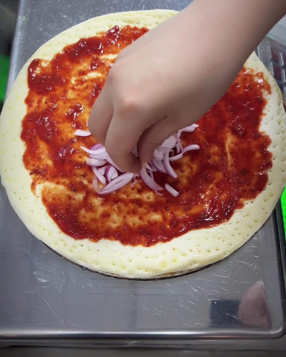 Rote Zwiebelstücke und eine dicke Schicht geriebener Mozzarella werden auf der Pizza verteilt.