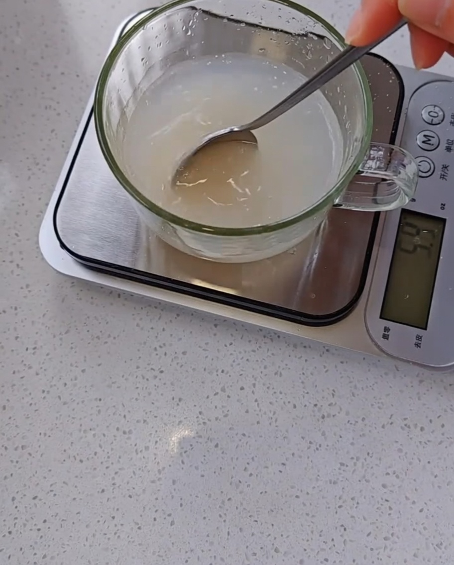 Eine Hand rührt mit einem Löffel eine trübe Mischung aus Hefe und Wasser in einem Messbecher auf einer digitalen Küchenwaage.