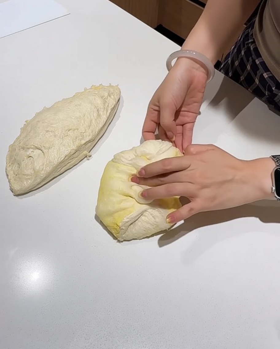 Hände formen ein Teigstück auf einer weißen Arbeitsfläche zu einer straffen Kugel.