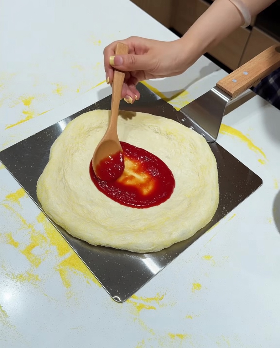 Ein Holzlöffel verteilt rote Tomatensauce auf dem Pizzateig auf einem Metallschieber.