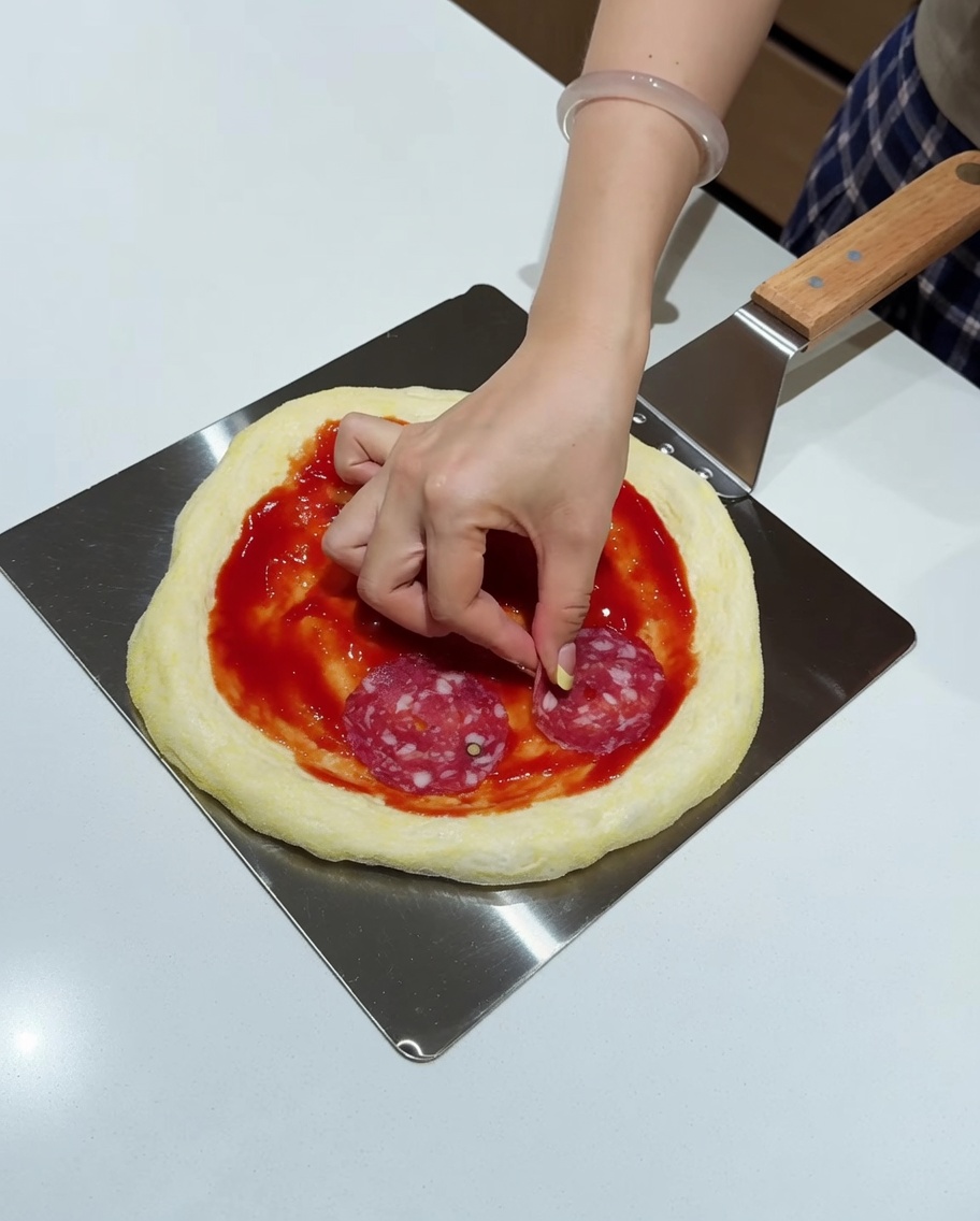 Hand platziert Salamischeiben auf einer belegten Pizza auf dem Schieber.