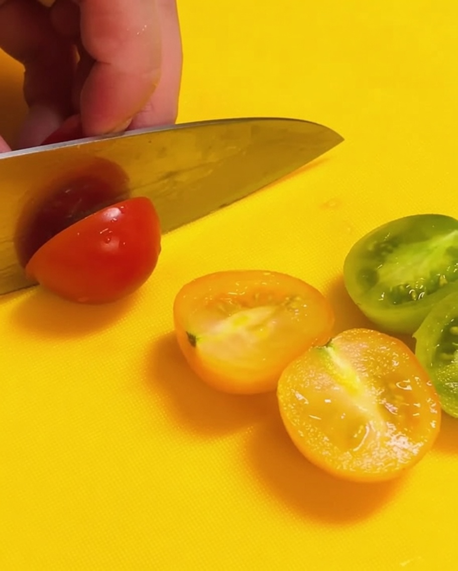 Ein Messer halbiert rote und grüne Kirschtomaten auf einem leuchtend gelben Schneidebrett.