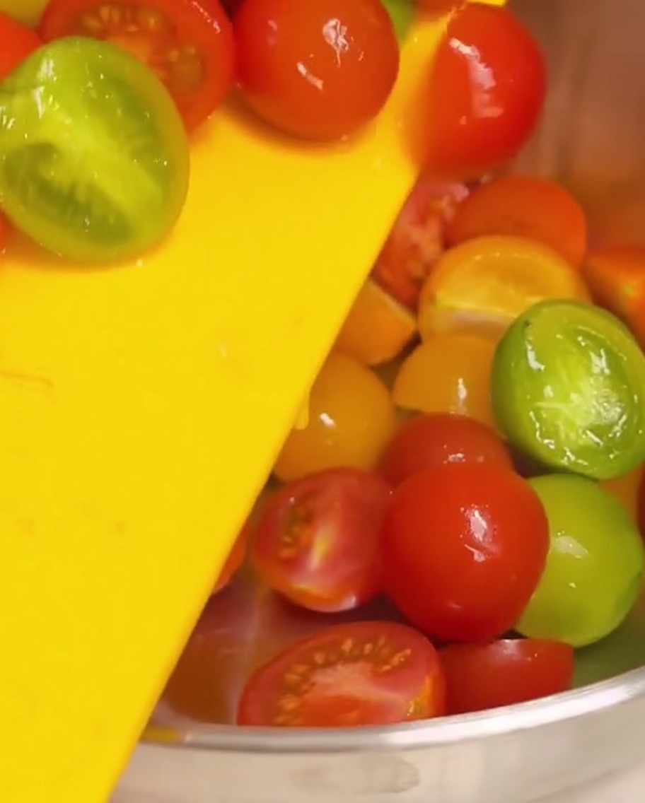Halbierte Kirschtomaten werden von einem gelben Schneidebrett in eine Edelstahlschüssel geschoben.