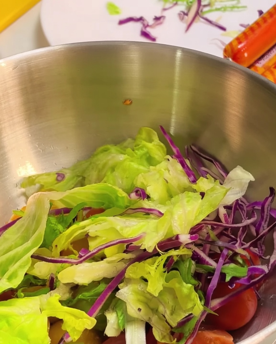 Frische grüne Salatblätter und zerkleinerter Rotkohl werden in eine Metallschüssel über die Kirschtomaten gegeben.