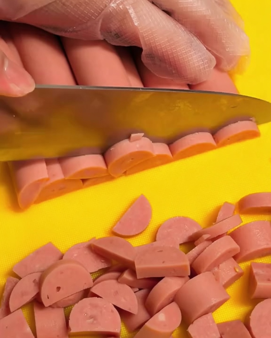 Eine Hand mit einem transparenten Handschuh schneidet rosa Schinkenwurst auf einem gelben Schneidebrett in kleine Halbmonde.