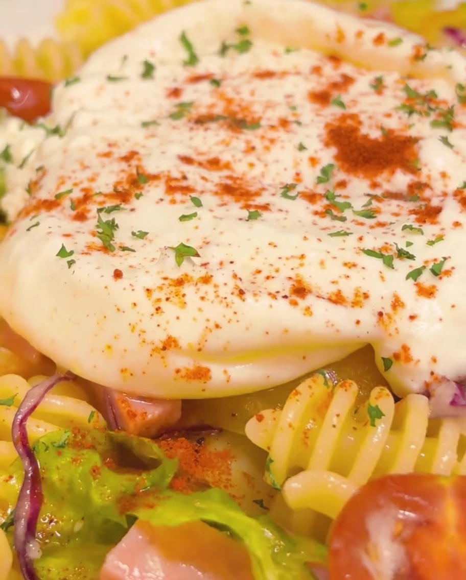 Nahaufnahme des fertigen kalten Nudelgerichts mit einem cremigen Burrata-Zentrum, garniert mit Petersilie und Paprika sowie einem finalen goldenen Spritzer Olivenöl.