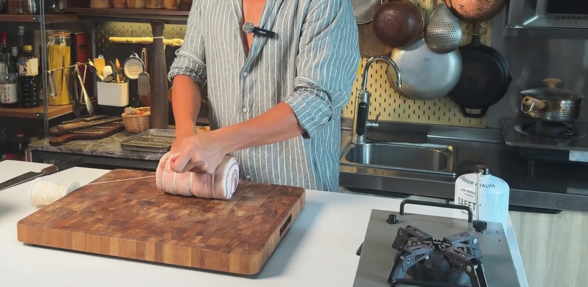 Ein gerollter roher Schweinebauch, der waagerecht mit Küchengarn auf einem Holzschneidebrett gebunden wird.