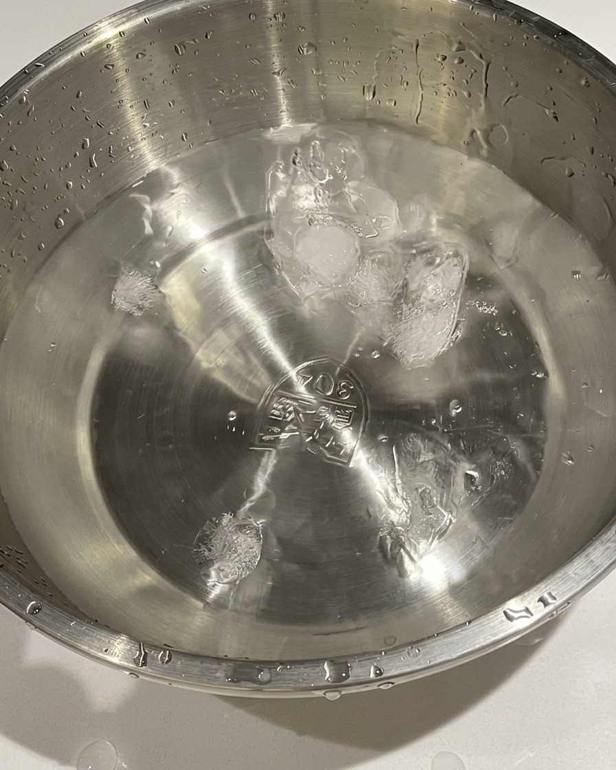 Gekochte Eier werden aus einem Topf in eine Metallschüssel mit kaltem Wasser und Eiswürfeln umgefüllt.