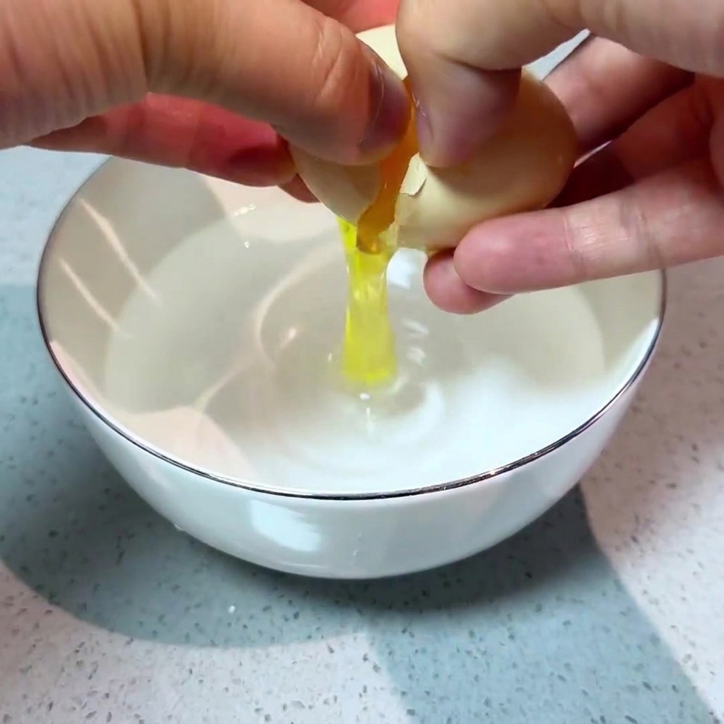 Zwei Hände schlagen vorsichtig ein frisches Ei in eine weiße Schüssel mit klarem Wasser auf.