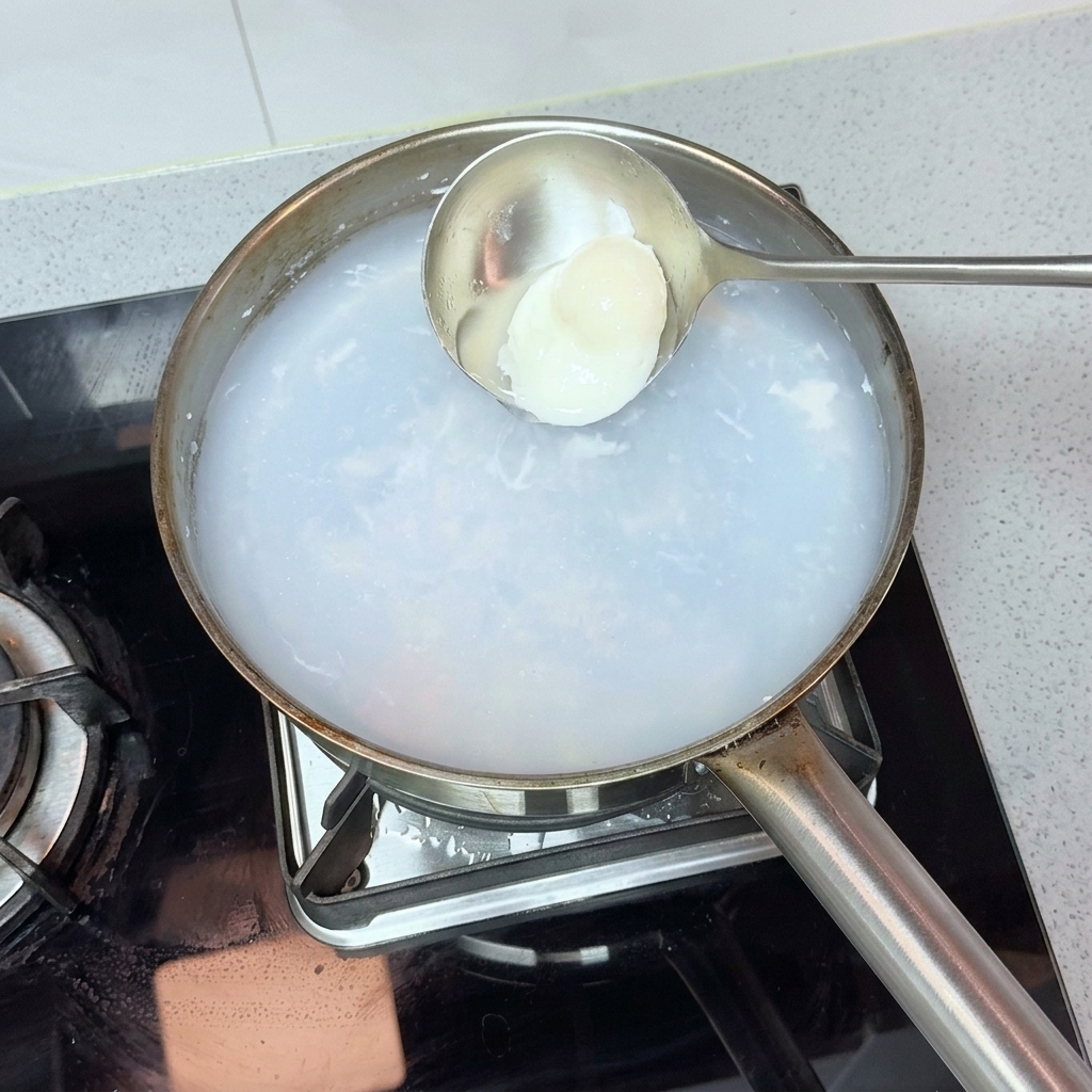 Eine Metallkelle schöpft vorsichtig ein perfekt pochiertes Ei aus einem Topf mit heißem, trübem Wasser.