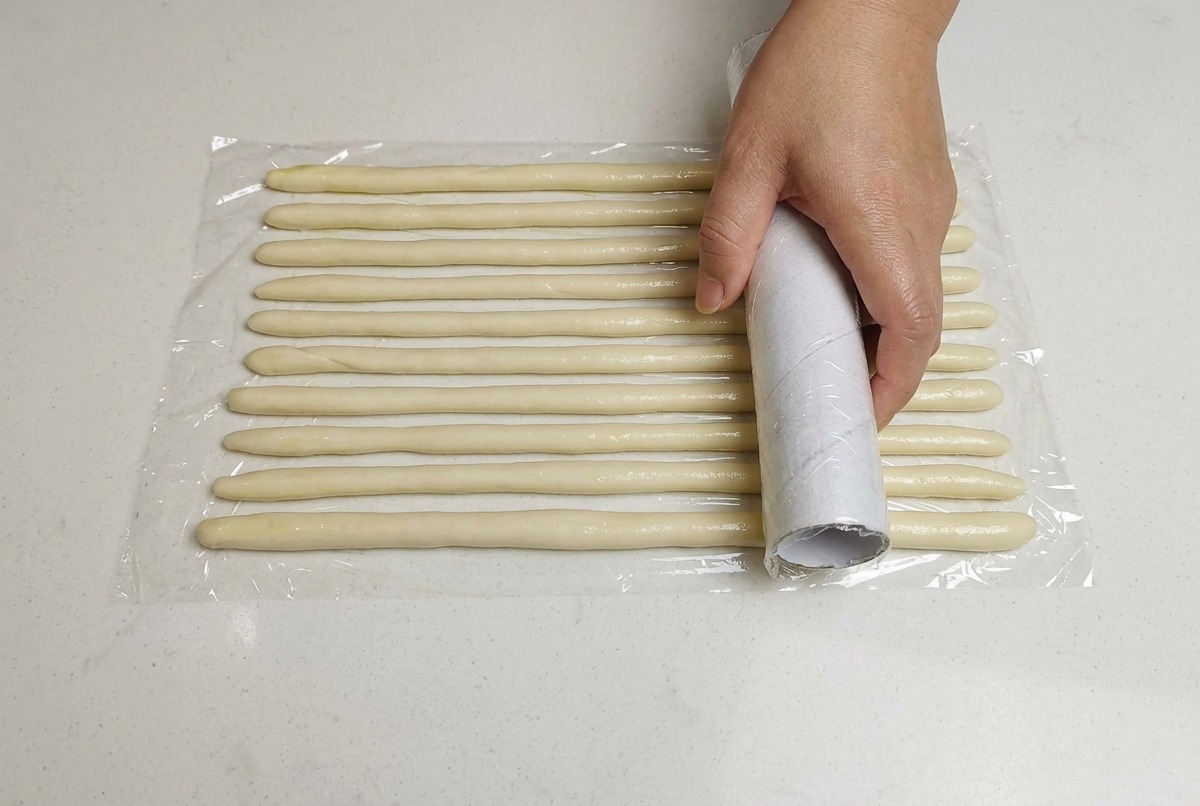 Eine Hand legt ein Blatt Frischhaltefolie über angeordnete, dünne Teigstränge auf einer weißen Oberfläche.