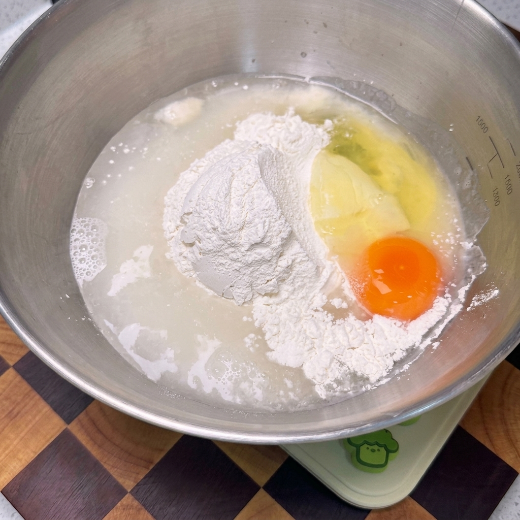 Ein Schneebesen mischt Mehl, Ei und Flüssigkeit in einer Edelstahlschüssel zu einem glatten, hellgelben Teig.