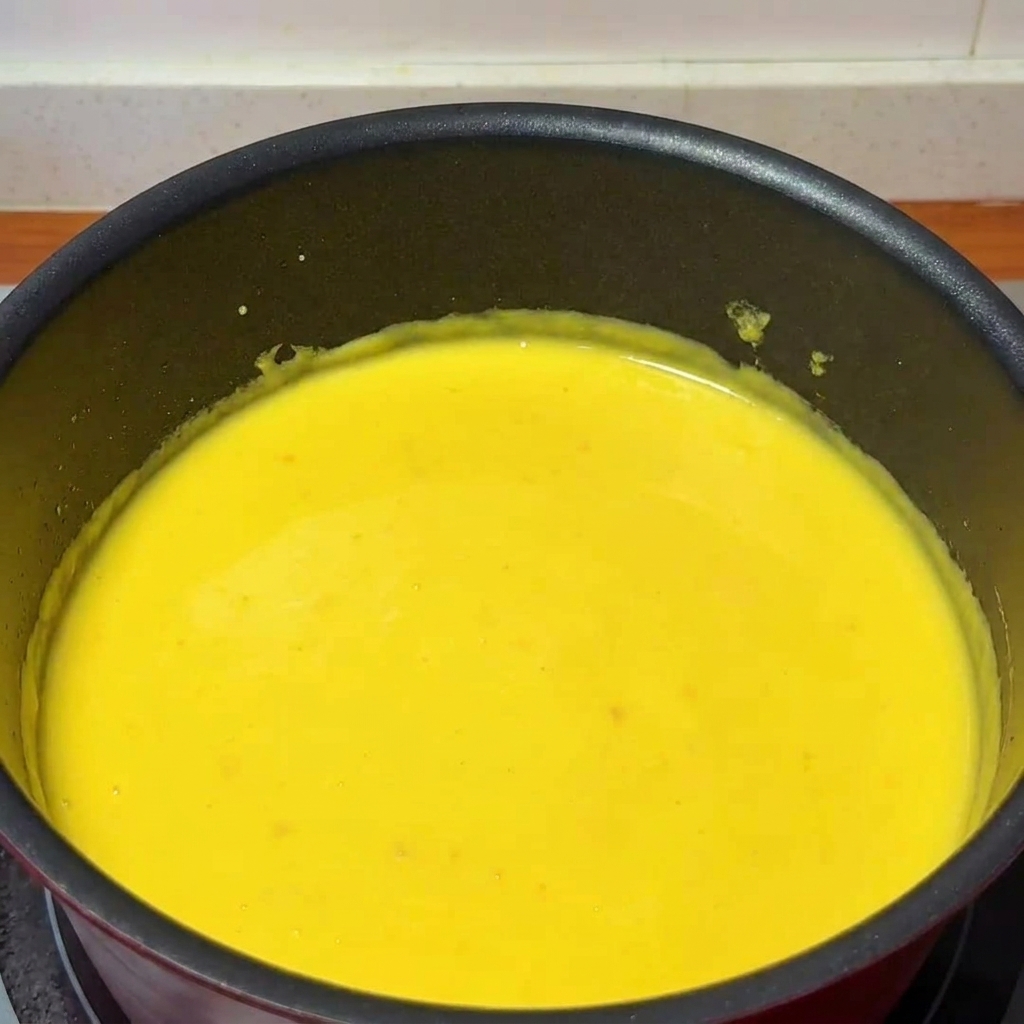 Eine Kelle gibt Yuzusaft in einen Topf mit dicker, cremiger Käsesauce, während mit einem Holzlöffel gerührt wird.