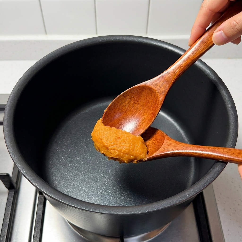 Ein Holzlöffel rührt Miso-Paste, Mirin, Zucker, Brühe und Yuzusaft in einem Topf.