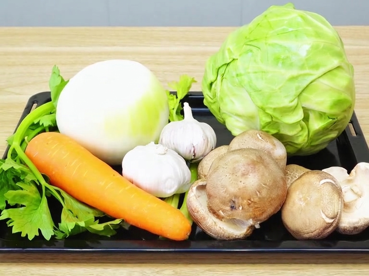 Eine Auswahl an frischem Gemüse, bestehend aus Zwiebeln, Knoblauch, Karotten, Sellerie, Shiitake und Kohl, ordentlich auf einem Holzbrett angerichtet.